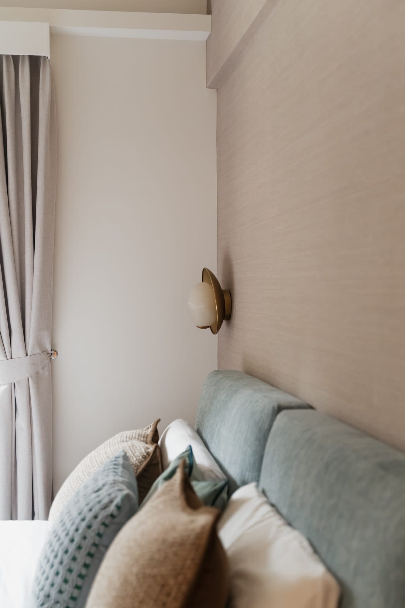 A bedroom corner: an upholstered headboard in soft blue-green against textured wallpaper, lit by a brass and opal glass wall light