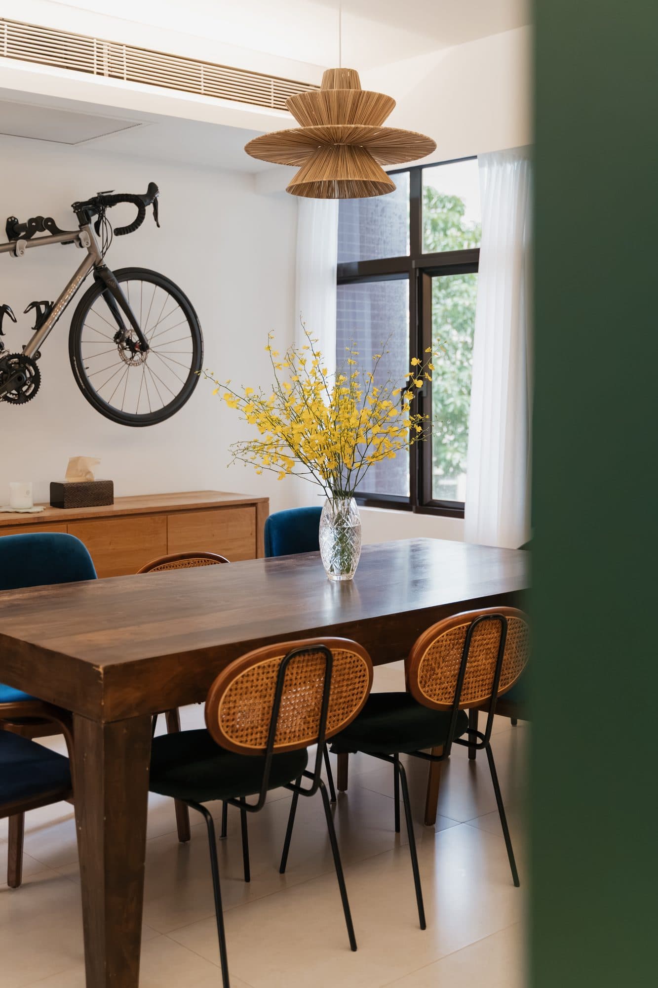 The dining room with a road bicycle wall-mounted above a low timber sideboard, beside a solid timber table and rattan-backed chairs