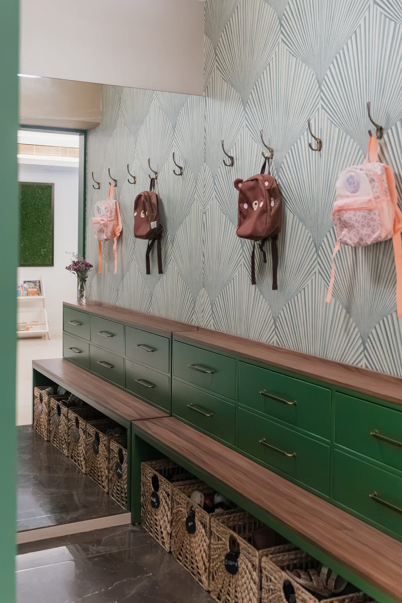 The entry: deep green joinery under a walnut bench top, woven baskets in the cubbies below, a row of brass hooks on a pale green fan-patterned paper