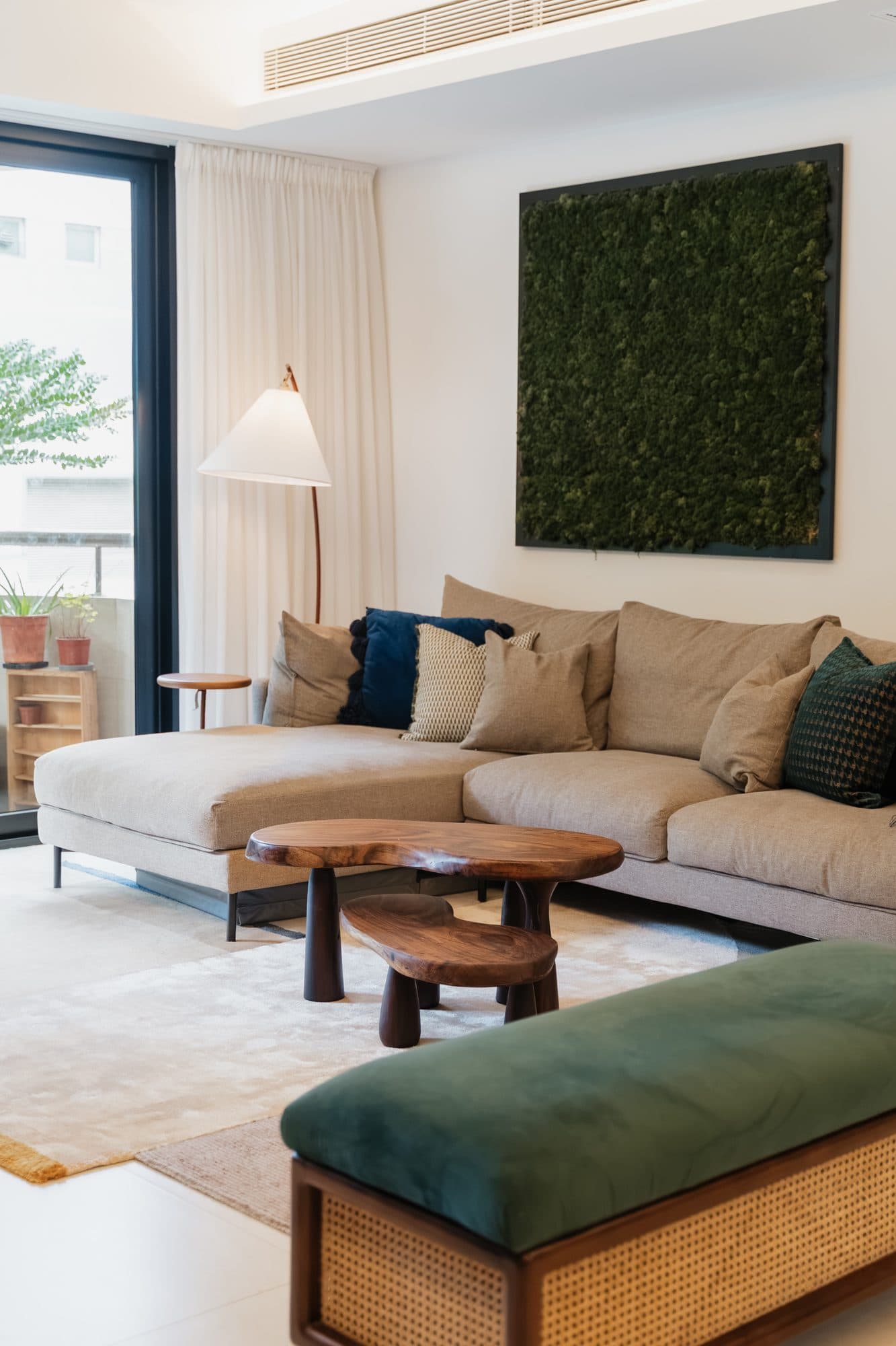 The living room from the window end: a low linen sectional under a framed moss panel, with an arc floor lamp and a caned bench in green velvet