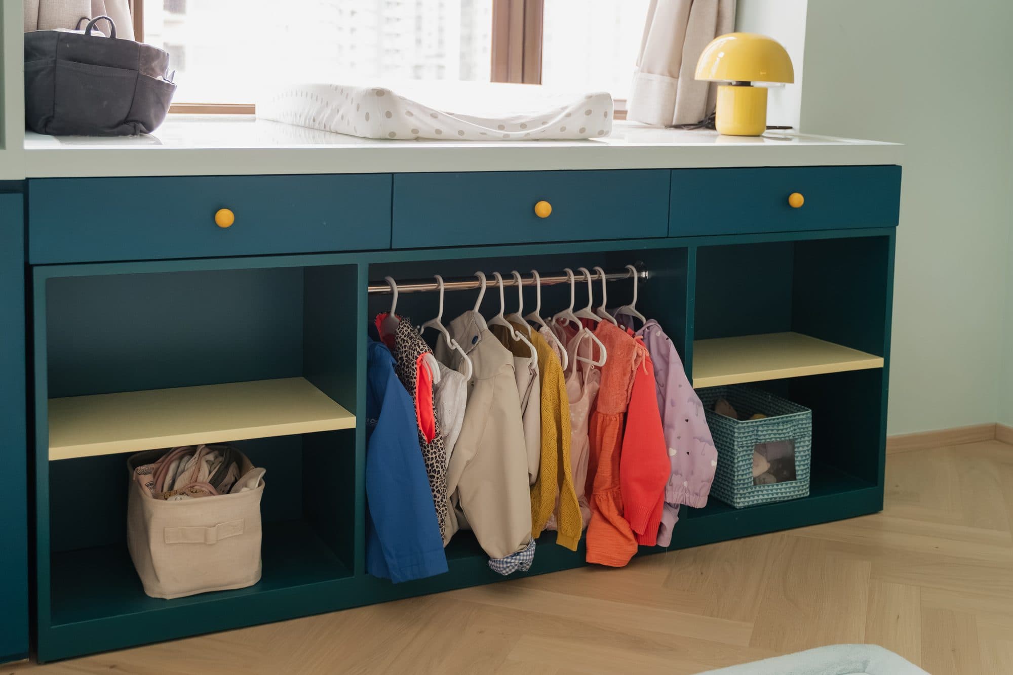 The Montessori wardrobe as built, in teal with yellow knobs: a low hanging rail of children's clothes with a shelved bay to either side