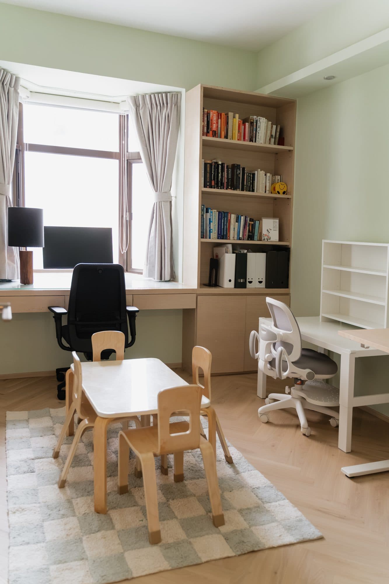 The study in soft green: a built-in desk running under the window beside a column of bookshelves, with a child-height table on a checkerboard rug
